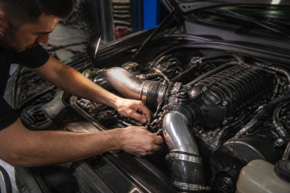 Technician examining intercooler piping and boost lines during supercharger installation inspection, hands checking connections in engine bay with proper lighting