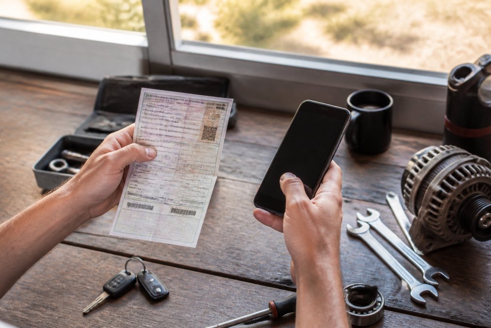 Close-up of hands holding a vehicle registration document and a smartphone displaying a parts compatibility search tool on screen, with natural light from a window
