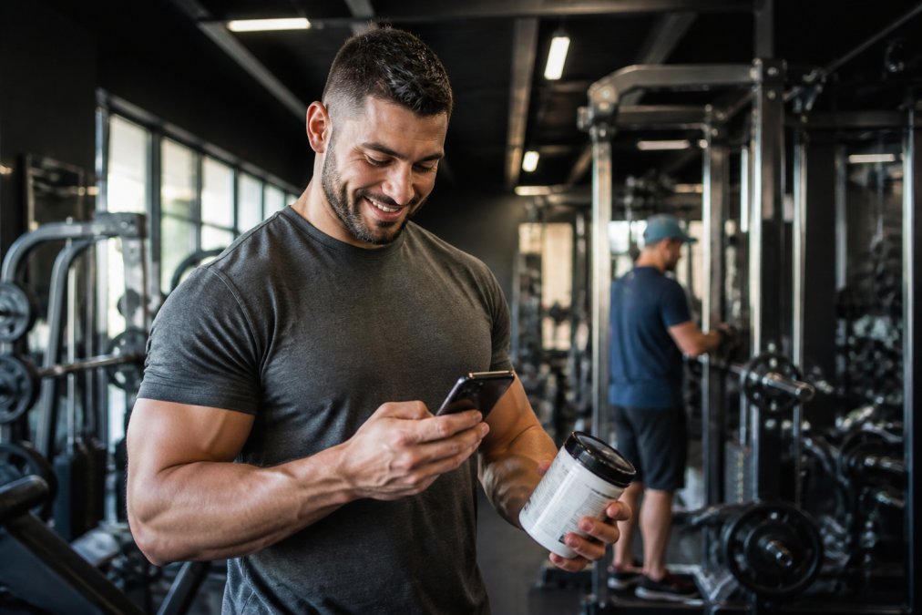 Athlete checking phone rewards dashboard while holding supplement bottle at gym, natural gym lighting with equipment in background