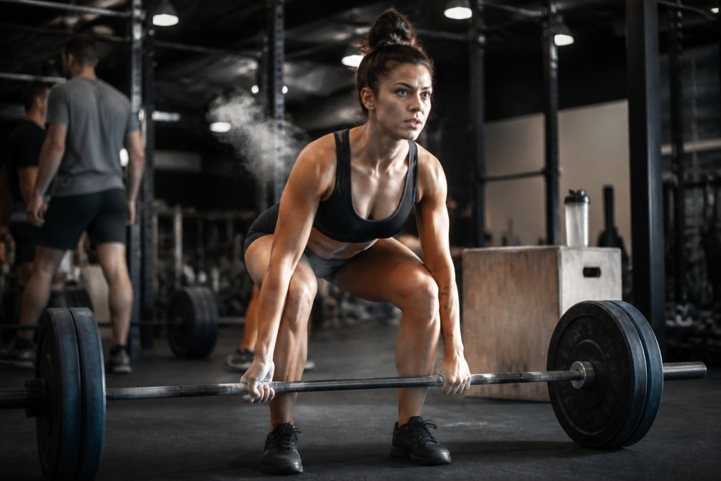 A CrossFit athlete mid-WOD in a functional fitness gym, gripping a loaded barbell with a plyometric box behind them, chalk dust hanging in the air under industrial lighting