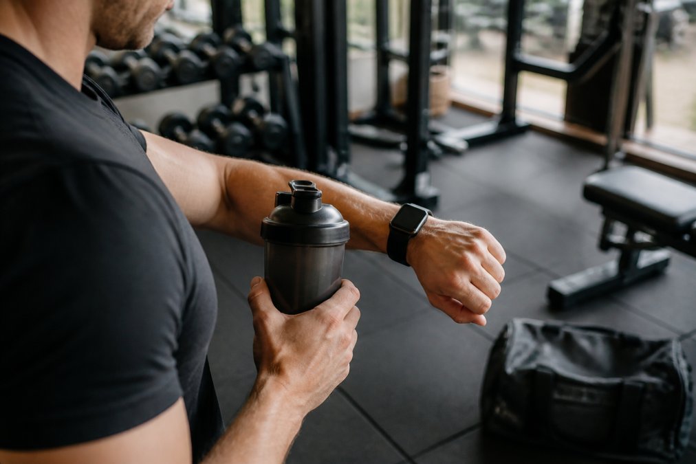 Close-up of hands holding pre-workout drink and checking time on smartwatch before heading to gym, athletic setting with gym equipment visible