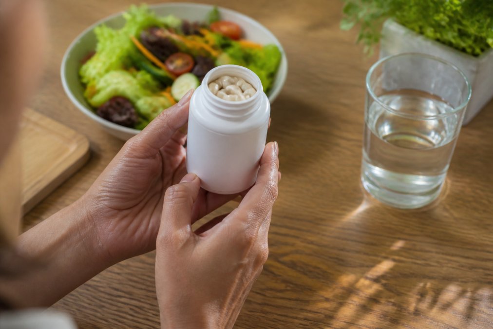Close-up of someone's hands holding a probiotic supplement bottle with a vibrant mixed green salad and a glass of filtered water visible on a wooden table in soft, warm afternoon lighting