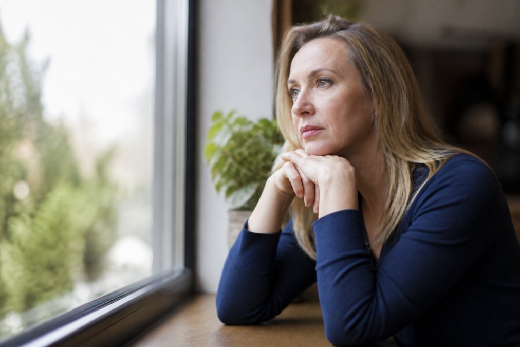Woman in her mid-40s sitting by a window with natural daylight, looking thoughtful with a subtle expression of contemplation, hands resting near her face, suggesting the emotional complexity of hormonal transition