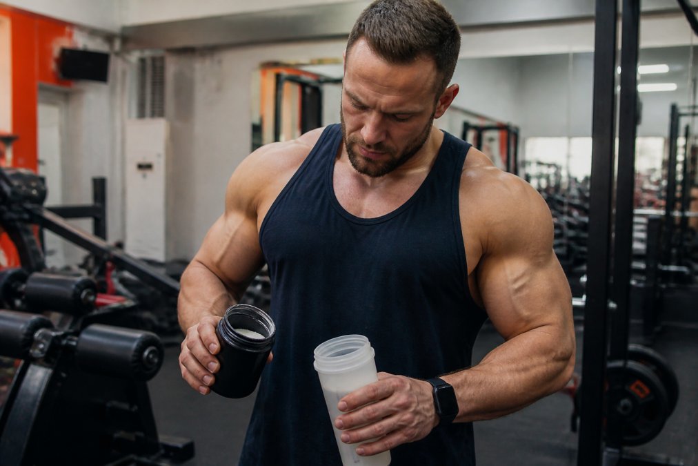 Athletic individual in modern gym setting holding supplement container and water bottle, preparing pre-workout routine with focused intensity, bright overhead lighting