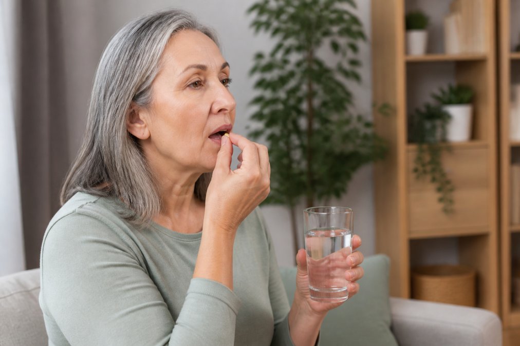A mature woman in her 50s taking a supplement capsule with a glass of water at home, looking calm and focused on her wellness routine