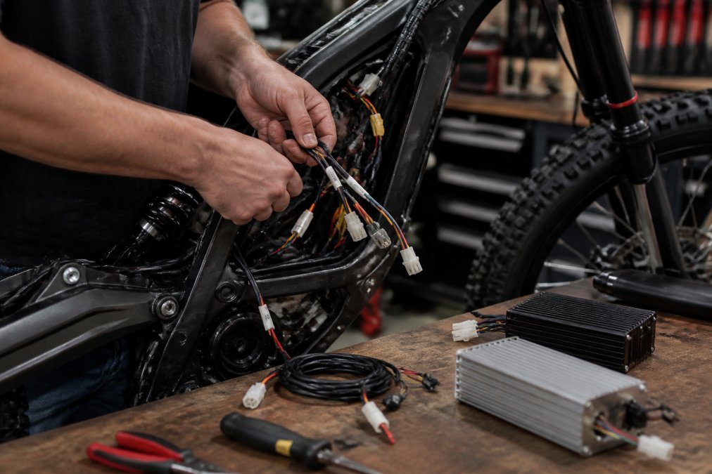 A mechanic's hands routing a labeled wiring harness into a Sur Ron Light Bee frame on a workbench, controller and tools laid out nearby