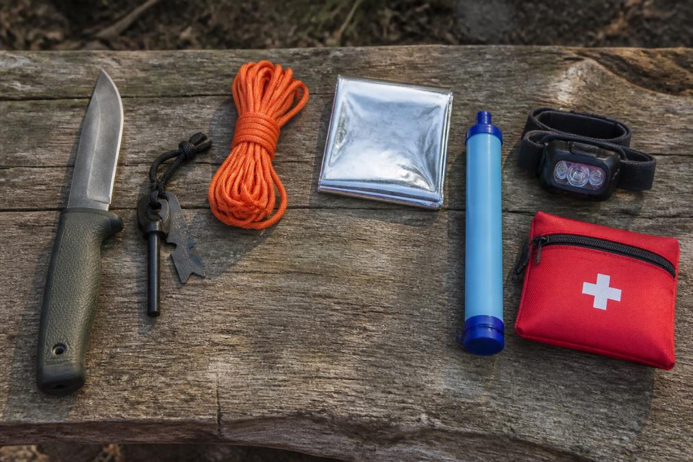 Flat lay of essential wilderness survival tools arranged on a weathered wooden surface in natural daylight: a fixed blade knife with polymer handle, magnesium fire starter, orange paracord coil, silver mylar thermal blanket, water filter straw, compact headlamp, and small red first aid pouch