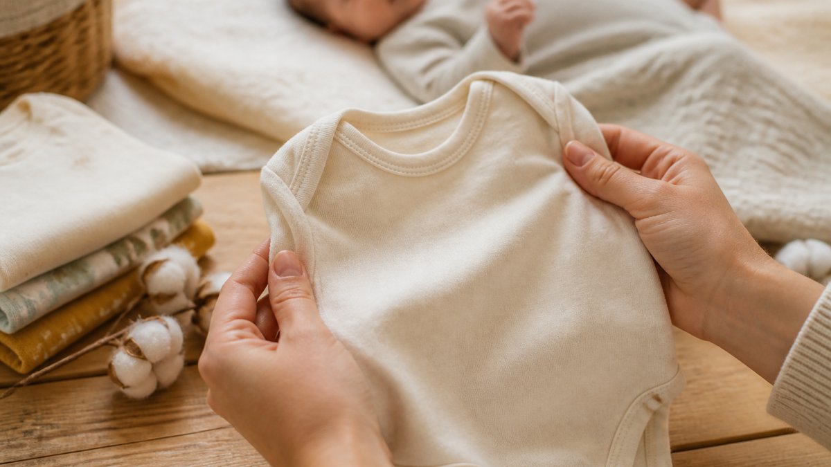 A parent's hands holding a soft organic cotton baby onesie against a natural wooden surface, with a blurred baby in the background