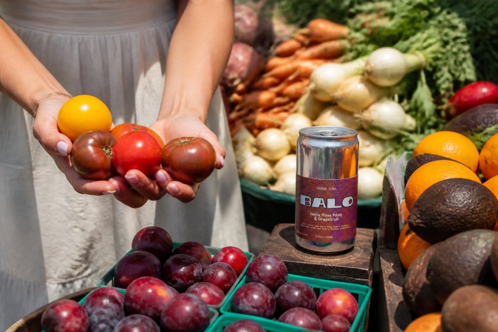Hands holding freshly harvested colorful produce at a farmers market stand with various fruits and vegetables displayed, showing the source of sustainable drink ingredients in bright natural light