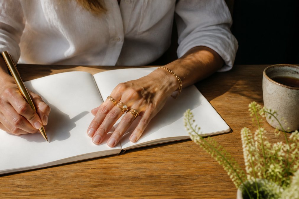 A woman's hands wearing stacked gold rings and a bracelet while writing in a journal at a sunlit wooden desk, showing the pieces in an everyday setting