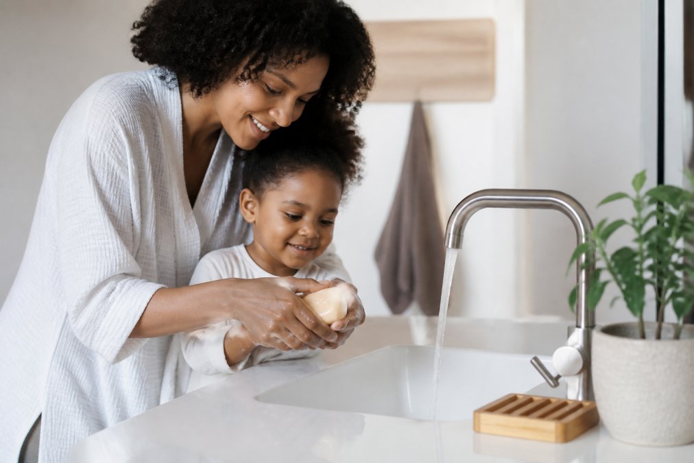 A mother helping her young child wash their hands with a natural beige bar of soap in a bright, clean bathroom, with a bamboo soap dish nearby