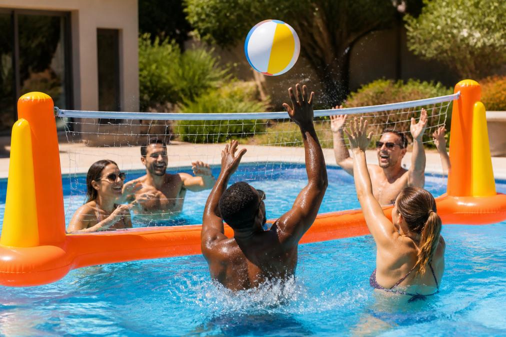 Group of adults laughing and competing over an inflatable volleyball net in a sunny backyard pool, beach ball mid-air, water splashing on both sides in warm afternoon light