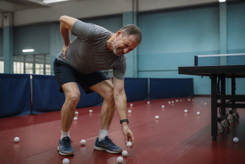 A table tennis player in his 40s grimacing while bending over to pick up scattered balls, holding his lower back with one hand, indoor club lighting