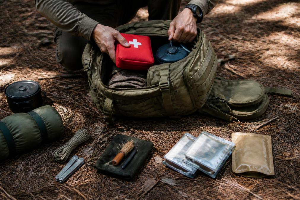 Hands methodically packing a large olive-drab tactical backpack with survival essentials including a red first aid kit, rolled hydration bladder, fire starter, and compact food rations spread out on dry pine needle-covered forest ground in afternoon light