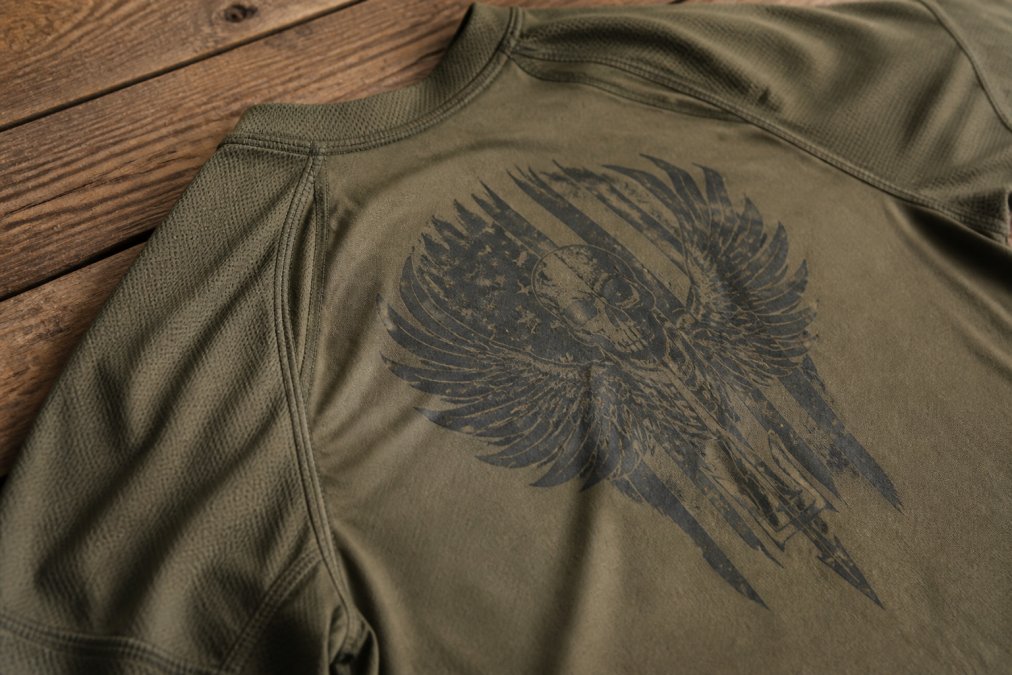 Close-up of a tactical shirt laid flat on a wooden surface showing fabric texture, reinforced stitching at the seams, and a military-inspired graphic print