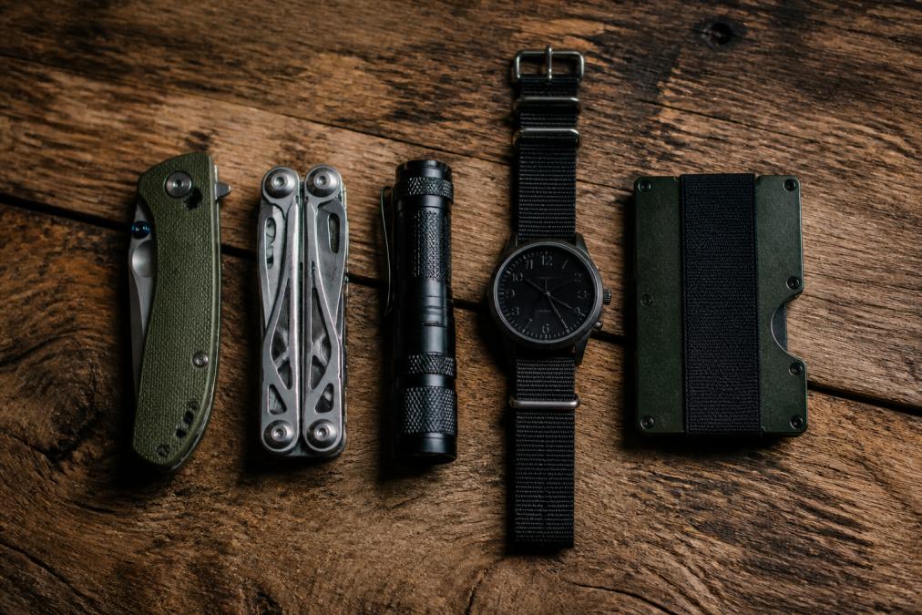 A flat lay photo of a complete everyday carry setup on a rugged wooden surface, featuring a folding knife, a multi-tool, a compact flashlight, a durable watch, and a slim cardholder wallet arranged neatly