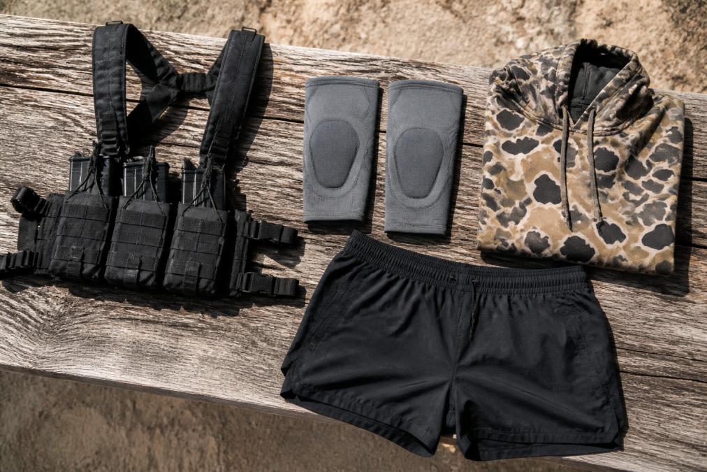 Flat lay of tactical gear on a weathered wooden bench: black chest rig with magazine pouches, gray knee sleeves, folded duck camo hoodie, and dark athletic shorts under harsh outdoor sunlight