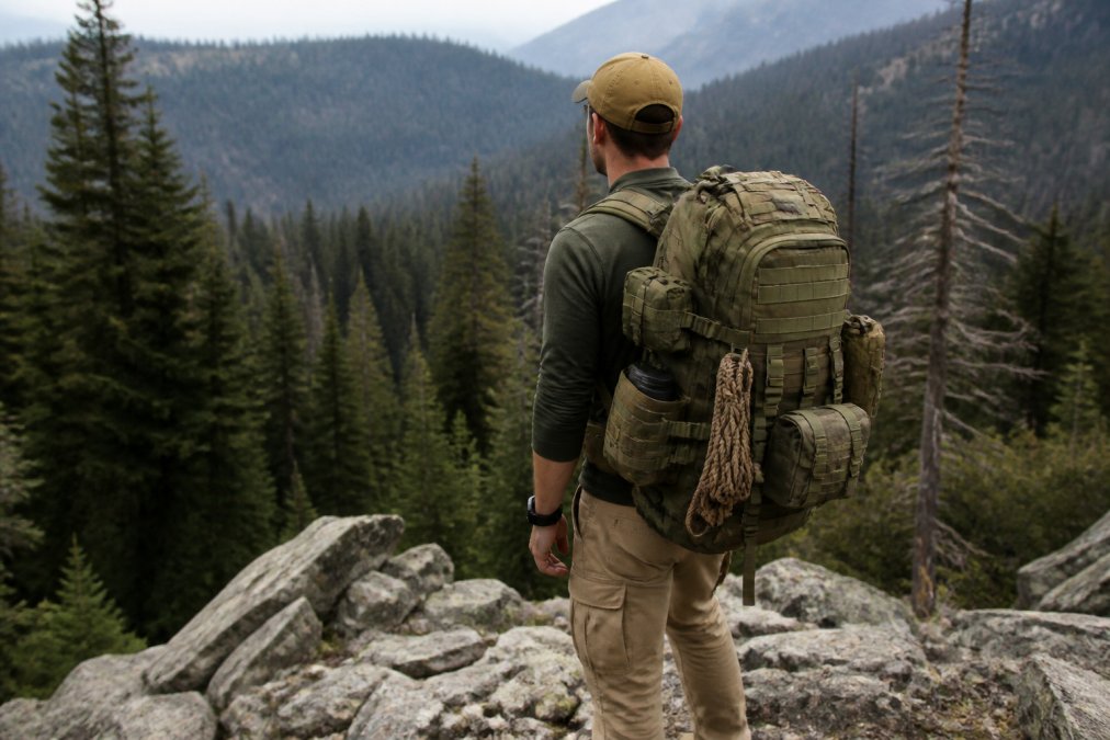 Hiker wearing a fully loaded tactical backpack with visible MOLLE webbing and modular pouches, standing on rocky mountain terrain with dense forest in the background