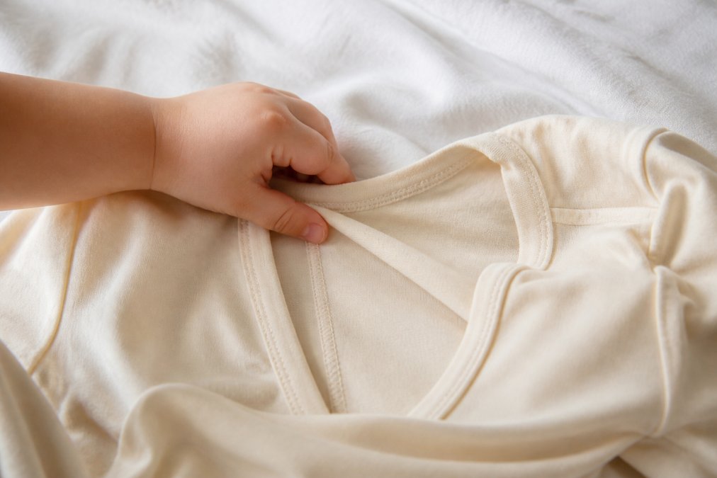 Close-up of a child's hand gently touching the interior of soft, cream-colored bamboo fabric pajamas laid flat on a white cotton surface, showing the smooth tagless neckline and flat seams in warm natural light