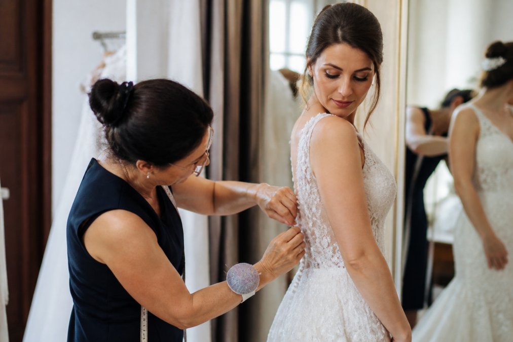 Professional seamstress carefully pinning and adjusting a wedding gown on a bride during fitting, with meticulous hand-stitching visible on delicate beaded fabric under soft studio lighting