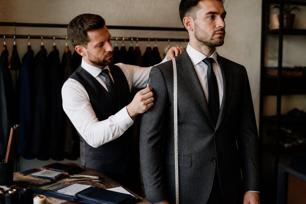 Professional tailor measuring client's jacket shoulders and chest with precision measuring tape during suit fitting consultation, with tailoring tools and fabric swatches visible on workbench