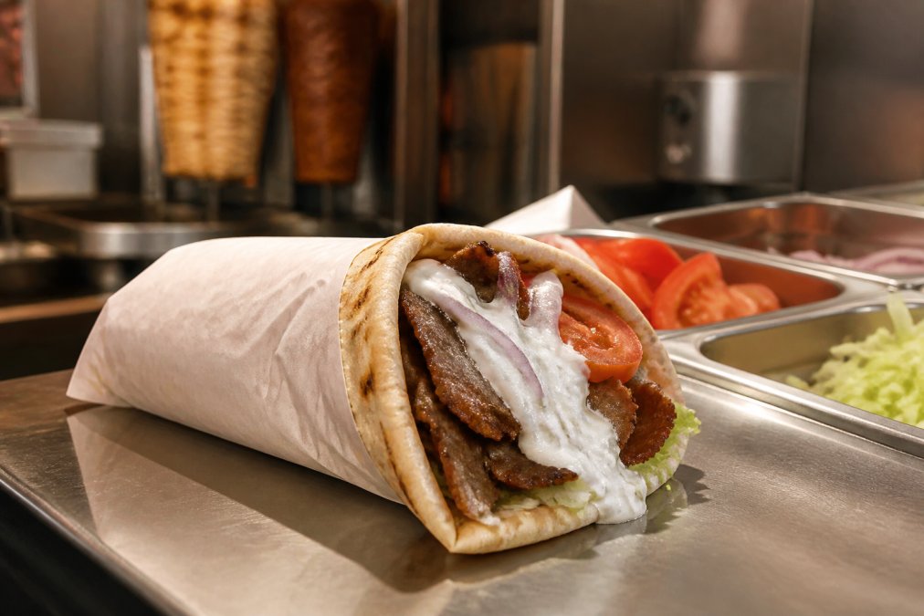 Close-up of a freshly prepared gyro with warm pita bread, visible layers of seasoned meat, fresh vegetables, and creamy tzatziki sauce, with a professional kitchen counter in soft warm lighting