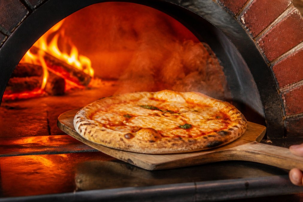 A wood-fired brick oven with a perfectly baked pizza being pulled out on a wooden peel, showing crispy charred crust and melted cheese, with steam rising and warm orange glow from inside the oven