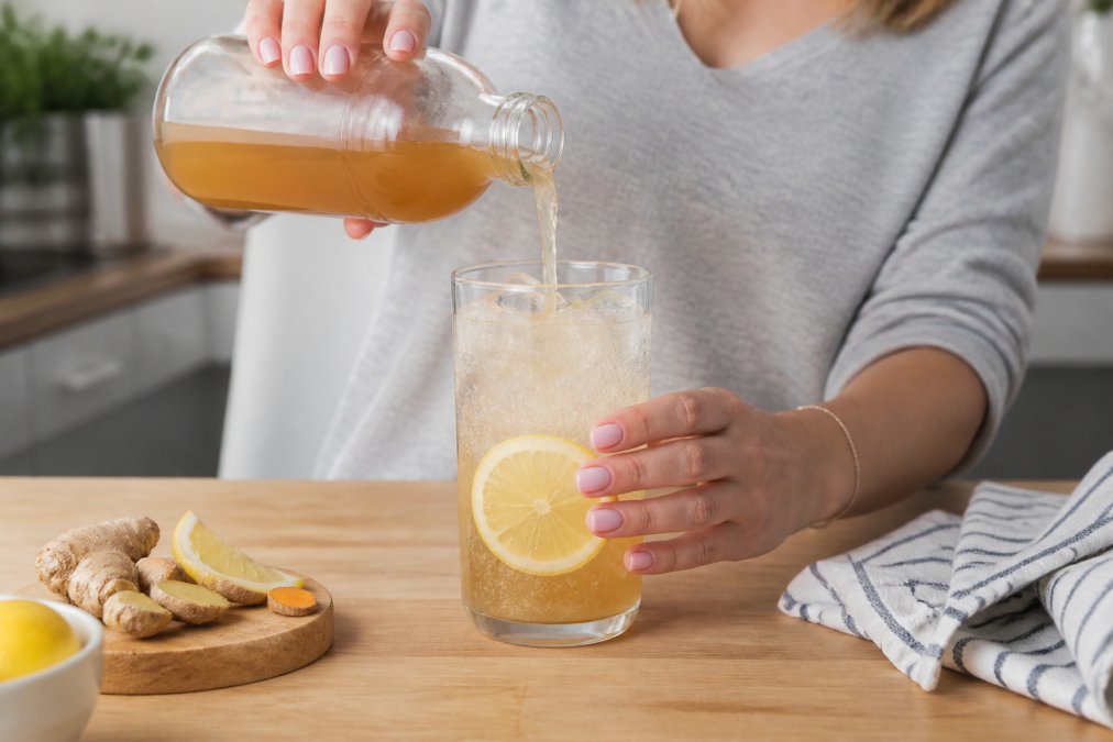 Person pouring Bite Me Tonic into a glass with sparkling water, ice, and a lemon wheel, showing the refreshing preparation ritual in a bright kitchen setting