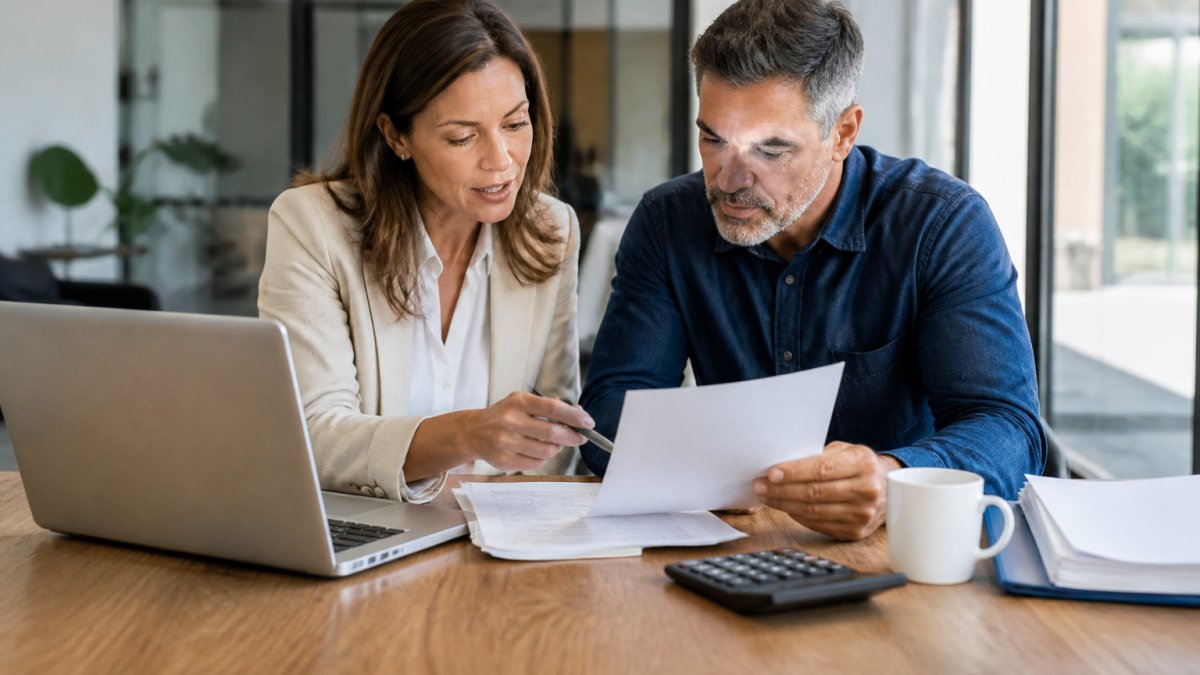 A tax professional and a small business owner reviewing financial documents together at a wooden desk, laptop open with spreadsheets, calculator and coffee cup nearby, bright natural light through office windows
