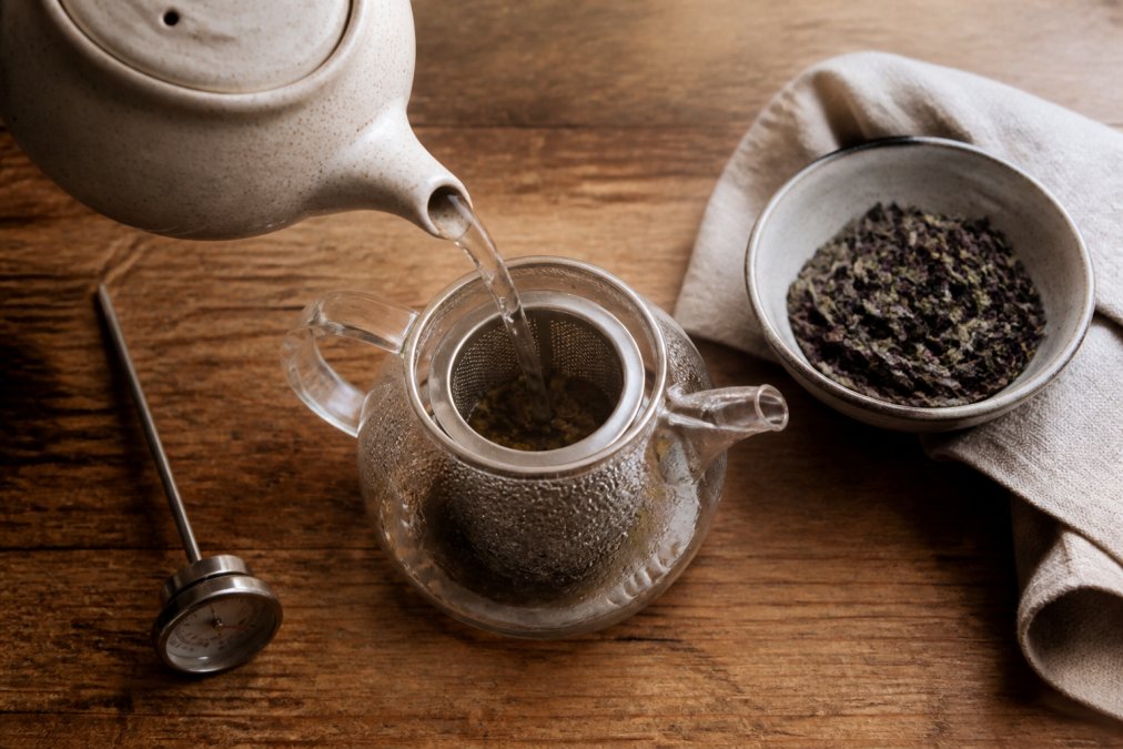 Overhead shot of a ceramic teapot pouring steaming water over loose leaf tea in a glass infuser, with a digital thermometer and a small bowl of dry tea leaves on a wooden kitchen counter