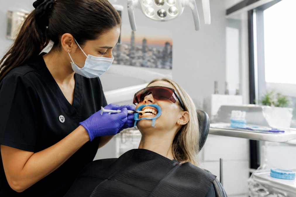 Dental professional carefully applying professional-grade whitening gel to patient's teeth with protective equipment in a modern clinical setting with bright overhead lighting