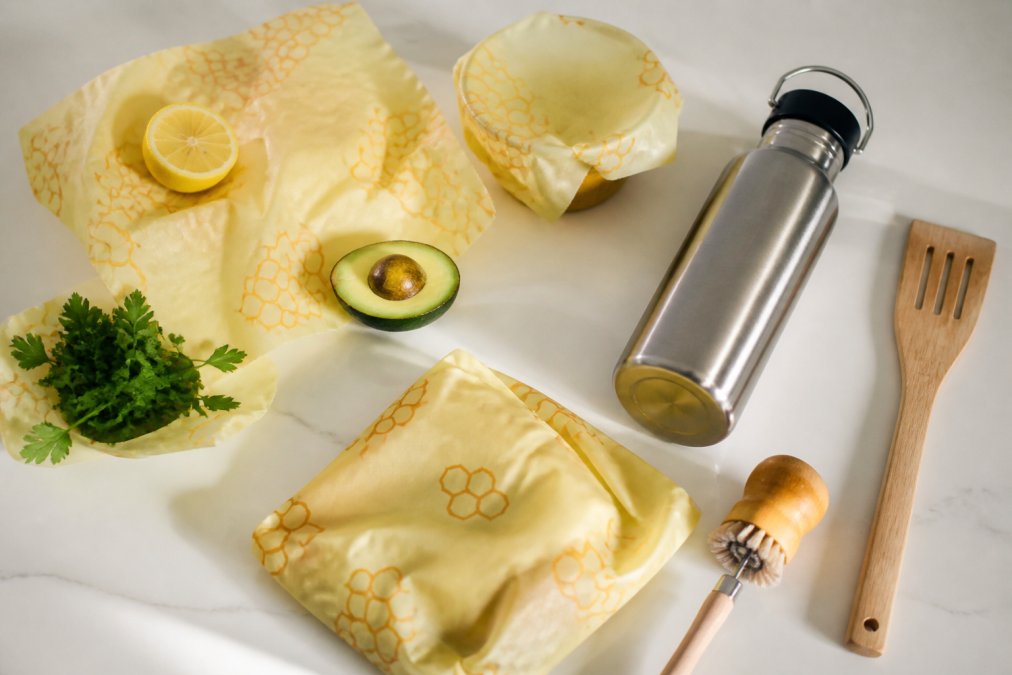 Flat-lay arrangement of household items including beeswax food wrap, kitchen tools, and a thermal bottle on a clean white surface with natural lighting