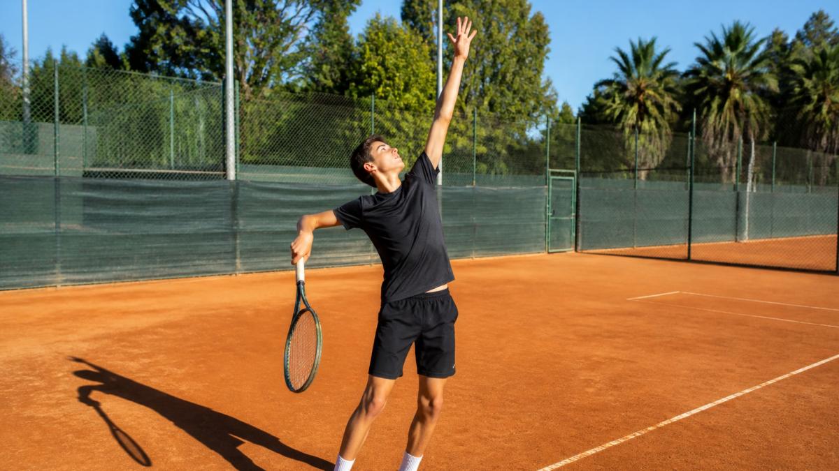 Young tennis player in trophy position on clay court showing proper shoulder rotation and racket drop before contact point, with morning sunlight casting shadows