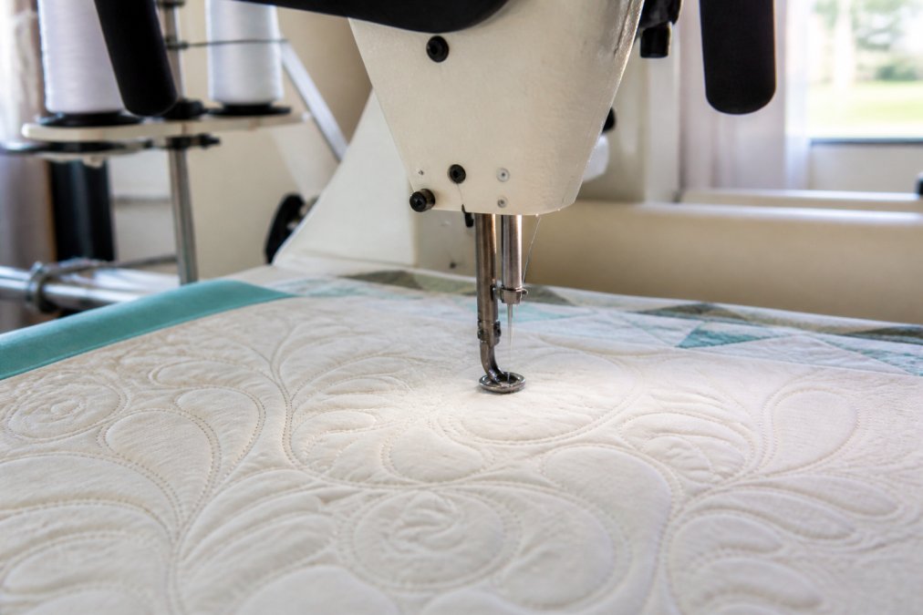 Close-up of a longarm quilting machine stitching with white Glide polyester thread, showing the smooth thread path and a quilt in progress under bright workshop lighting