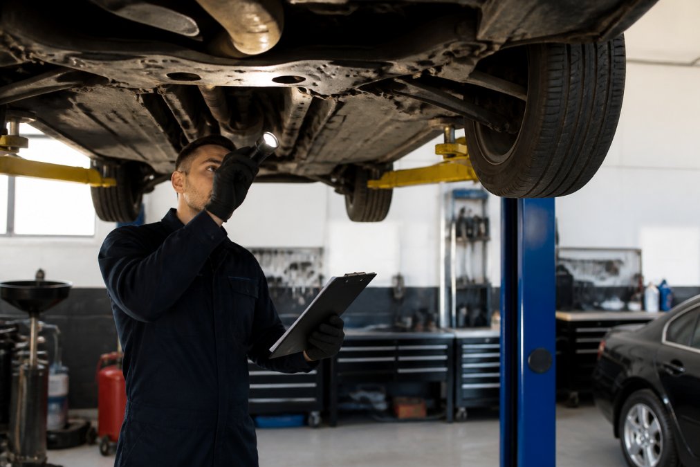 Professional examining the underside of a used sedan on a lift with a flashlight and clipboard, checking suspension and brake components in a well-lit service bay