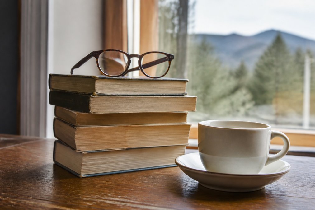 A stack of well-worn crime novels on a wooden table beside a cup of tea, with reading glasses resting on top and soft natural light from a window