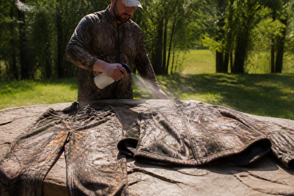 Hunter spraying permethrin treatment onto hunting jacket and pants laid out on a flat surface outdoors, with gear drying in sunlight before the hunt