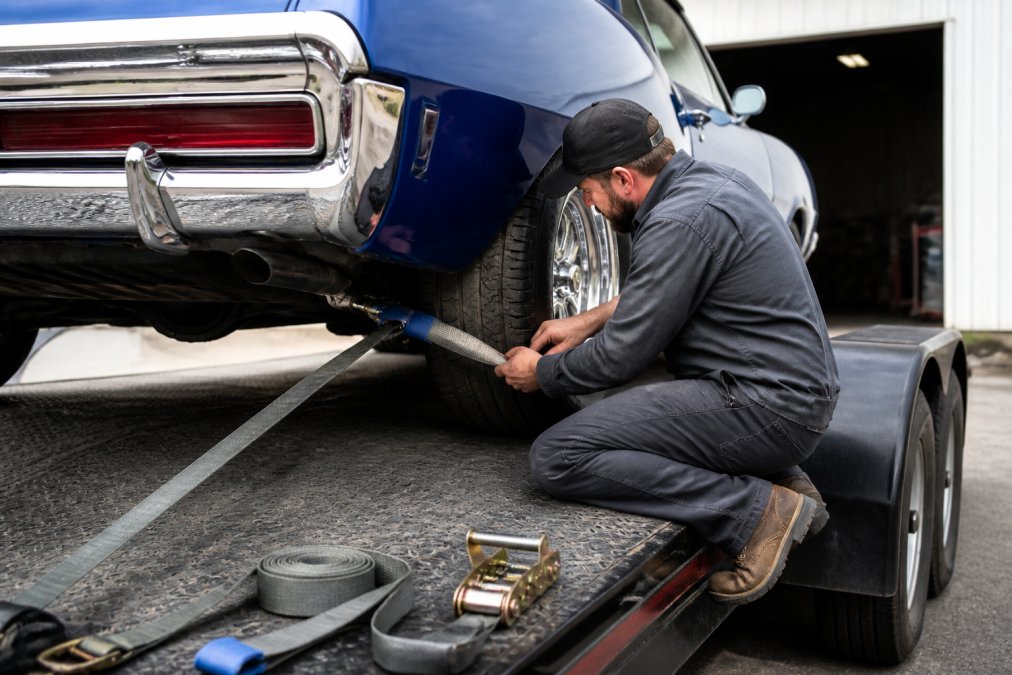 Person in work clothes securing a classic car to trailer tie down points using a soft loop and ratchet strap