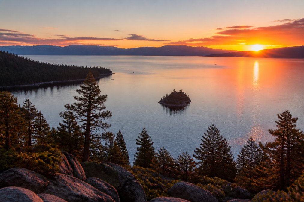 Golden hour light illuminating the Sierra Nevada mountains across Lake Tahoe, with the sun descending toward the horizon and casting warm orange and pink reflections across the calm water surface