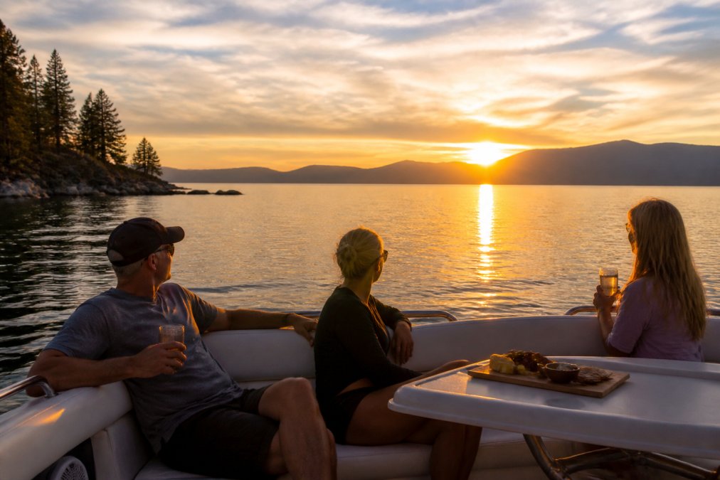 Small group of passengers relaxing on the deck of a private boat during golden hour, with the sun descending toward the Sierra Nevada mountains and warm light reflecting across the calm lake water