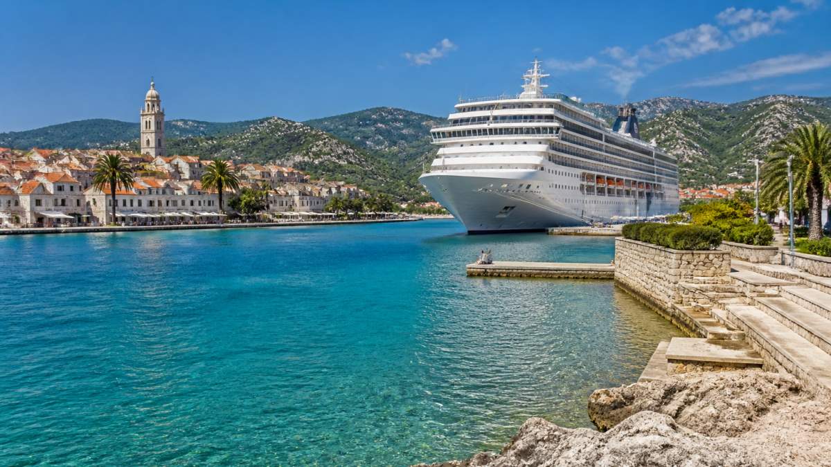Sunny Mediterranean coastal port with turquoise water, white stone buildings, and a large cruise ship docked at the harbor during bright daylight