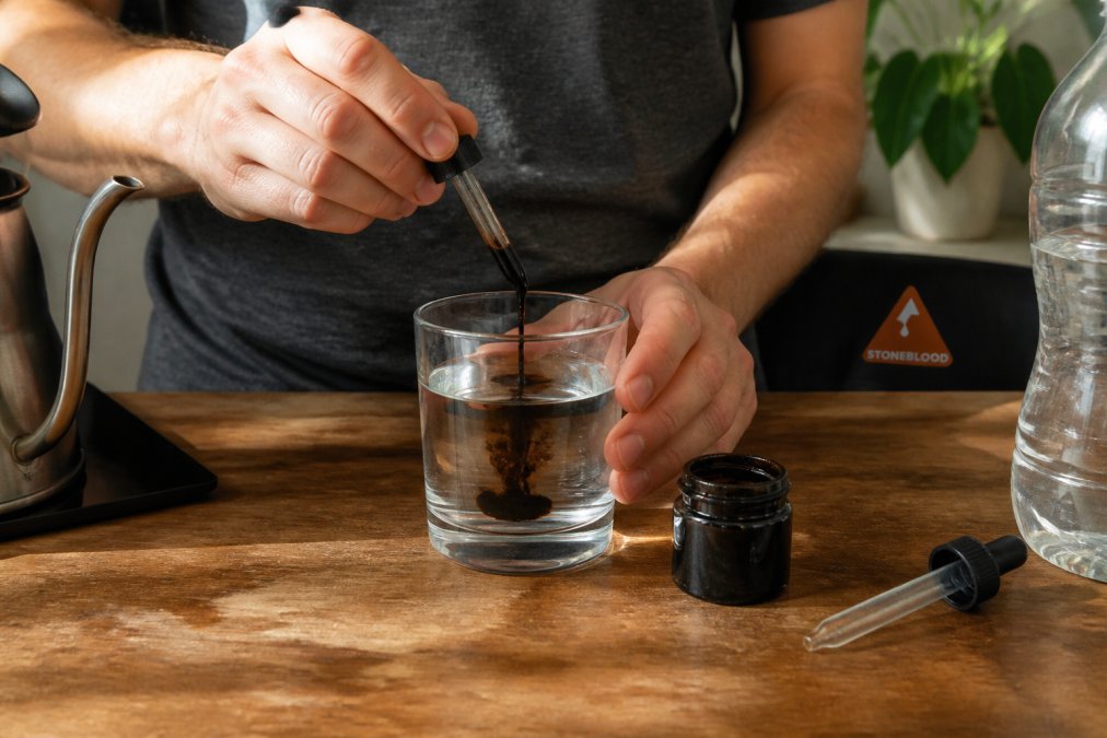Close-up of hands mixing dark shilajit resin into a glass of warm water on a wooden kitchen counter, with a small jar of resin and a measuring dropper nearby