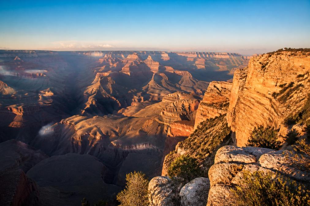 Sweeping aerial view of the Grand Canyon South Rim during golden hour with warm light illuminating layered canyon walls, clear blue sky overhead, and natural rock formations in sharp detail