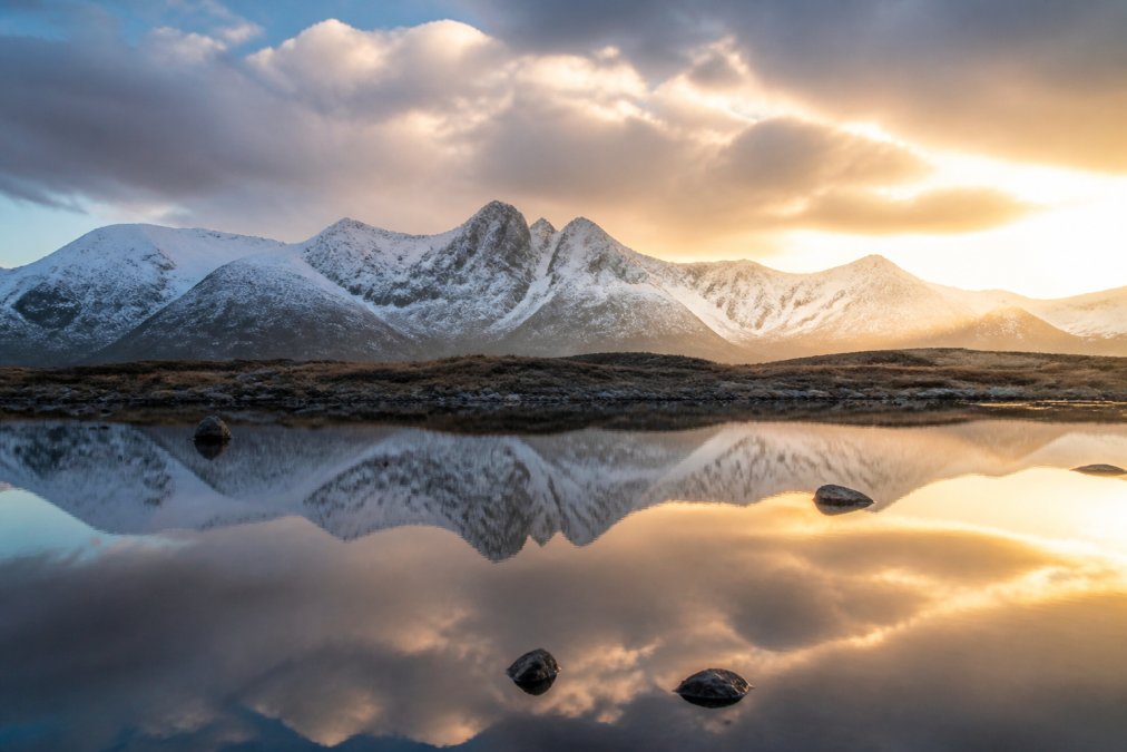 Dramatic landscape of snow-capped Cuillin peaks reflected in still water with golden hour light breaking through clouds