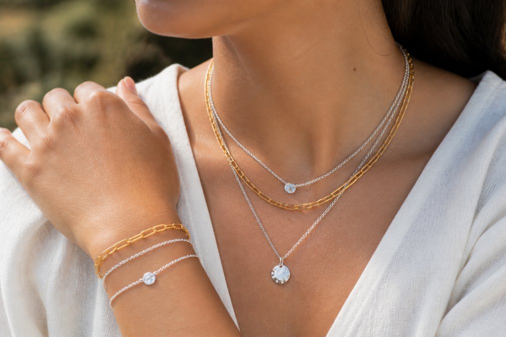 Close-up of delicate layered sterling silver and gold vermeil necklaces and bracelets worn on a woman's neck and wrist, showing how chains of varying lengths cascade together in natural daylight, demonstrating minimalist everyday elegance