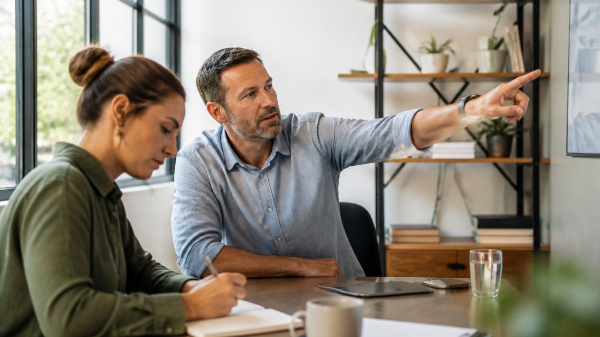A project manager pointing at a live project dashboard on a large monitor while a colleague takes notes in a bright agency office