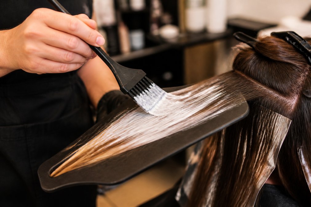 Close-up of a professional colorist's hand holding a feather bristle brush loaded with lightener, mid-stroke on a section of hair resting on a balayage paddle, warm salon lighting with blurred color station in the background