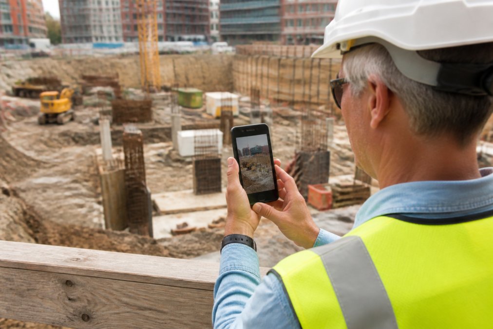 Field inspector using a mobile device to capture and annotate site photos at a construction site, with GPS coordinates and timestamp visible on the screen, natural outdoor lighting