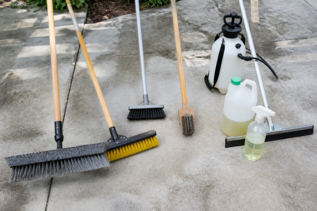 Organized collection of manual driveway cleaning tools laid out on concrete surface including push brooms with stiff bristles, wire brushes, floor squeegees, spray bottles, and containers of eco-friendly cleaning solutions arranged in natural daylight