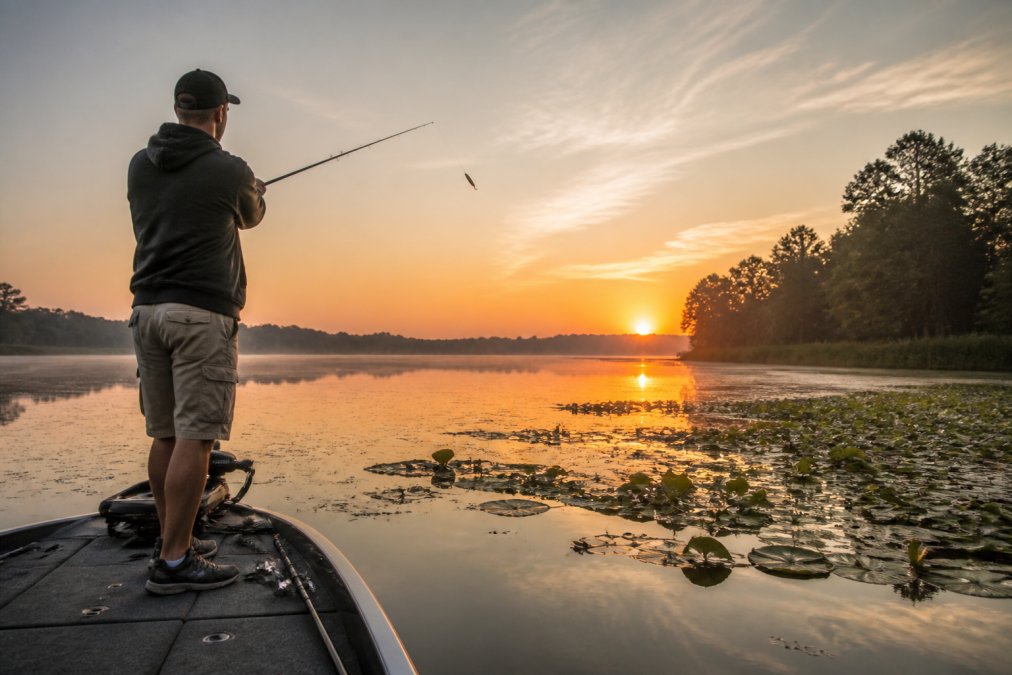 Angler casting a topwater frog lure toward lily pads at sunrise with golden light reflecting on calm water